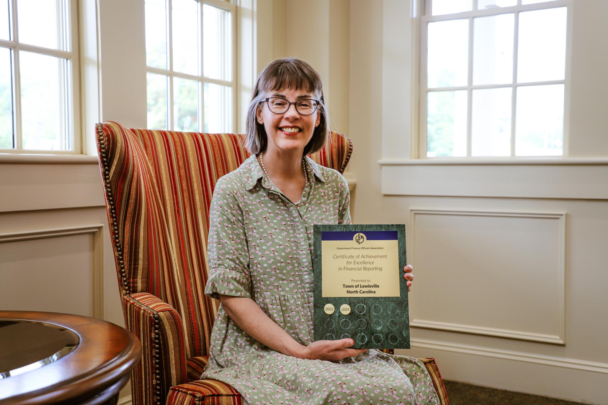 Pam Orrell poses with the Certificate of Achievement for Excellence in Financial Reporting, sitting on an armchair by a white wall with windows.