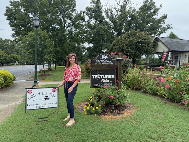 Textures Salon owner Diana Mitchell poses with the Yard of the Week sign in front of the salon. A beautiful green yard with colorful landscaping surrounds the business' sign and landscaping.