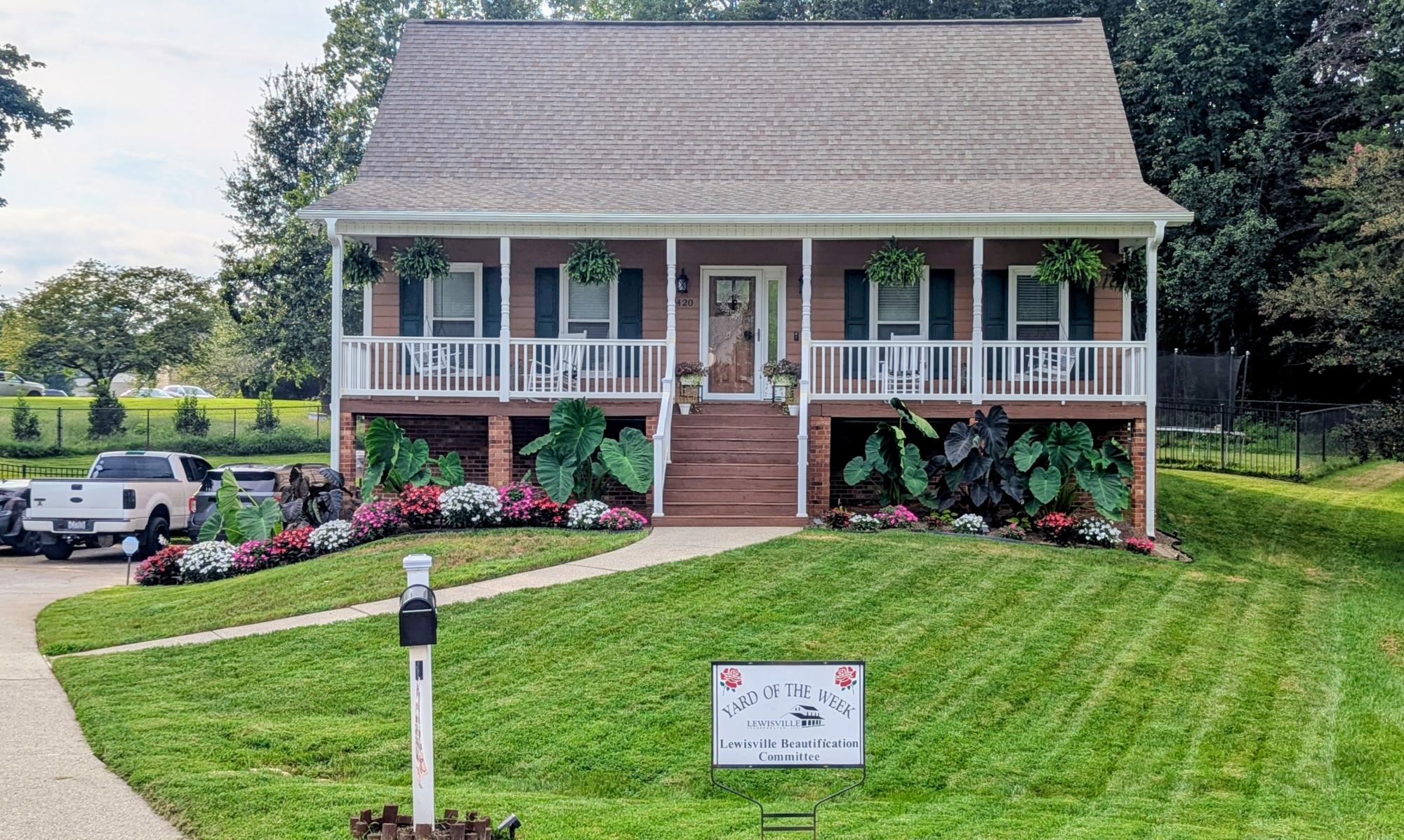 A stunning tan home has a large front porch with white railings and hanging greenery. Elephant ears and colorful flowers line the porch.
