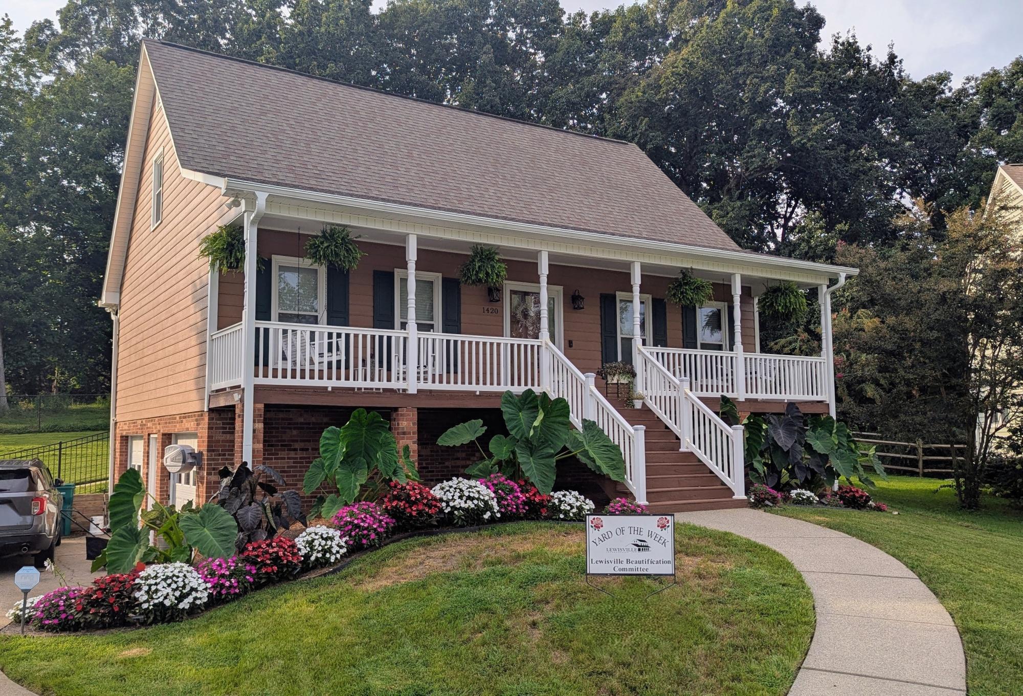 A gorgeous tan home is decorated with hanging greenery on the large front porch and colorful flowers along the bottom of the house