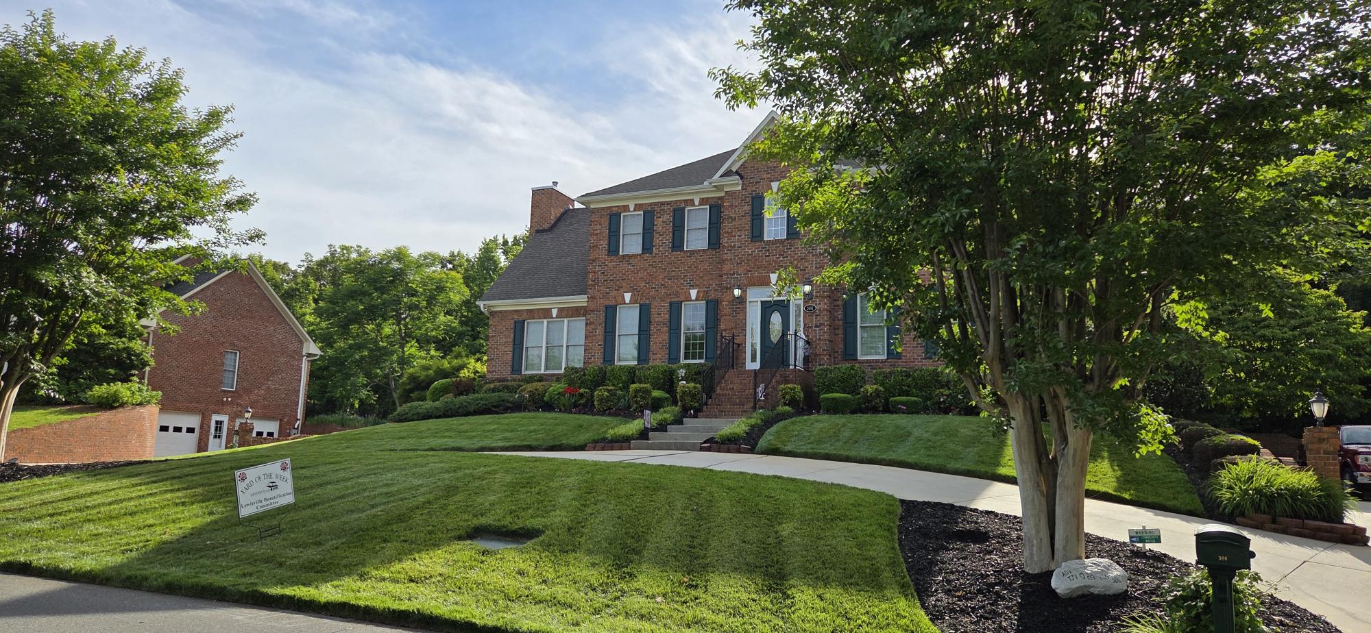 A beautiful brick home with a sweeping sidewalk and manicured front lawn on a bit of a hill has a "Yard of the Week" sign.