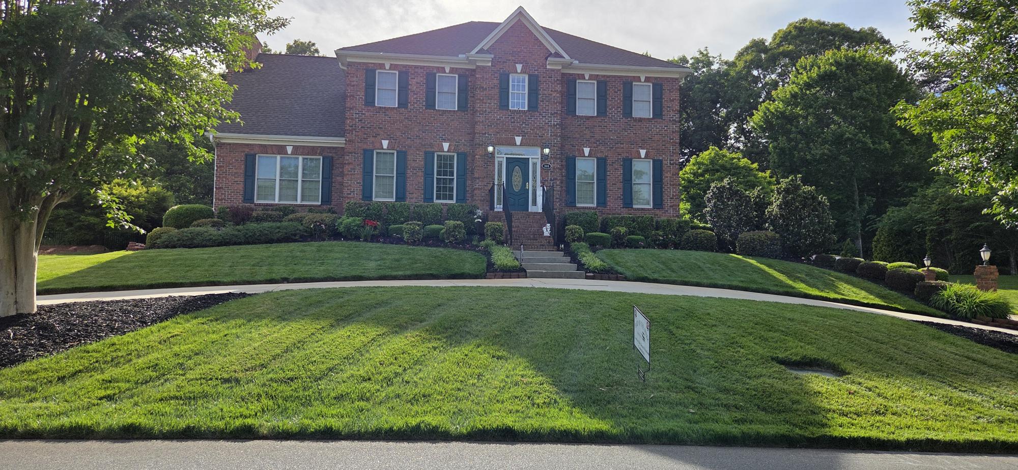 The lawn on the hill in front of a beautiful brick home is perfectly manicured. The neatly cared for bushes in front of the home are stunning. Large trees surround the home on either side.